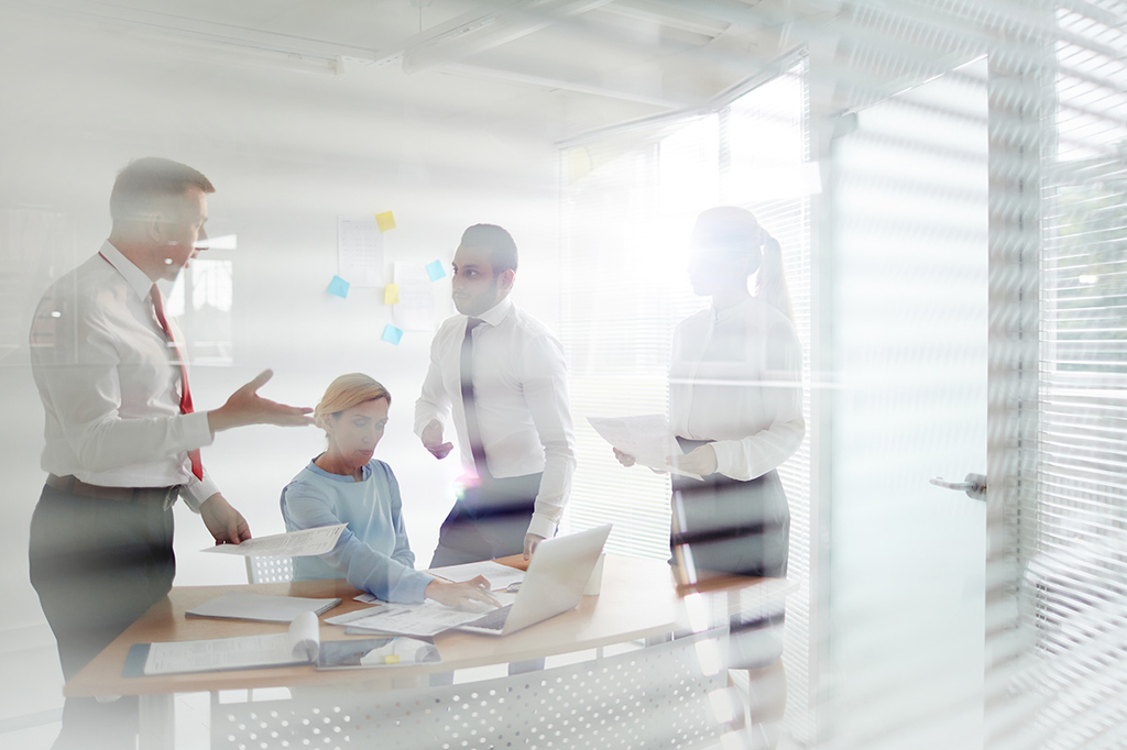 Blick durch eine Scheibe auf ein Büro, in dem Geschäftsleute mit Laptop und Dokumenten diskutieren.