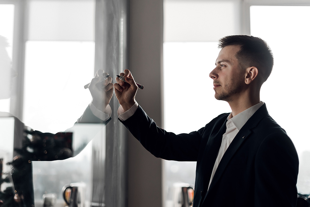 Junger Mann im schwarzen Anzug macht Notizen an einem Whiteboard.