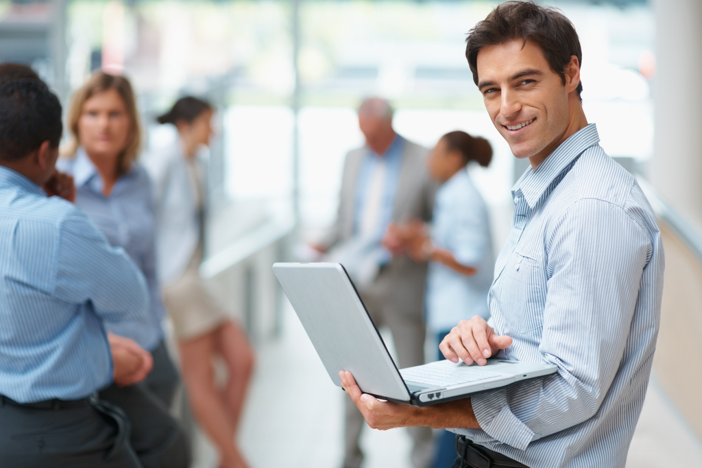 Junger Mann im Hemd hält einen offenen Laptop und lächelt in die Kamera.
